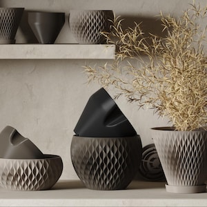 May include: A collection of modern, geometric planters in various shades of gray and black. The planters feature a textured, diamond-patterned design. One planter holds a dried plant with thin, beige leaves. The planters are displayed on a shelf.