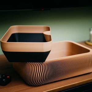 May include: Two-tone storage containers. The top container is a square shape with a tan rim and black interior. The bottom container is a rectangular shape with a corrugated, tan exterior. Both containers are on a wooden surface.