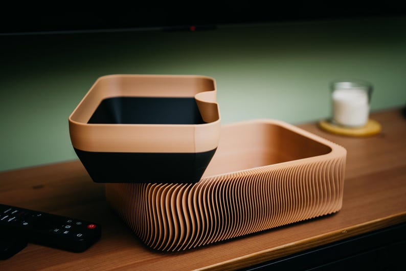 May include: Two-tone storage containers. The top container is a square shape with a tan rim and black interior. The bottom container is a rectangular shape with a corrugated, tan exterior. Both containers are on a wooden surface.