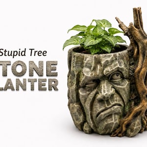 May include: A gray stone planter shaped like a human face with a grumpy expression, featuring a brown tree branch attached to the side. The text 'Stupid Tree STONE PLANTER' is printed to the left. A small green plant grows from the top opening.