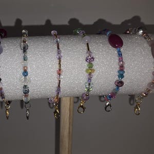 May include: Six beaded bracelets in various colors, including purple, pink, blue, and clear. The bracelets are displayed on a white cylindrical stand. Each bracelet features a different design with a clasp for easy wear.