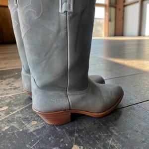 Puede incluir: Un par de botas vaqueras Dingo de color gris a media pantorrilla, con punta redondeada y tacón de madera. Las botas presentan patrones de costuras blancas decorativas en la caña y el nombre de la marca Dingo grabado en el talón lateral.