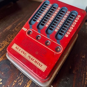 May include: A vintage red metal adding machine with four vertical sliding columns featuring numbers one through nine. The device includes a horizontal display window for the total and text that reads ADDING MACHINE PATENT 2243884.