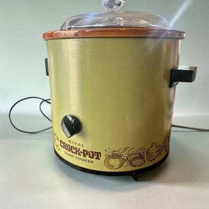 Può includere: Una slow cooker Rival Crock-Pot vintage con corpo cilindrico verde chiaro, bordo marrone e coperchio in vetro trasparente. La parte anteriore presenta una manopola di controllo nera e illustrazioni grafiche marroni di utensili con il testo Rival Crock-Pot Slow Cooker.