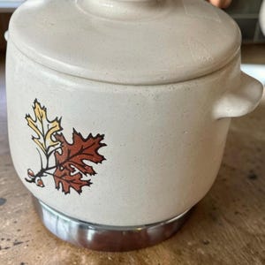 Op de afbeelding: Een crèmekleurige keramische pot met bijpassend deksel en twee handvatten aan de zijkanten. De buitenkant heeft een gespikkelde afwerking en een grafische illustratie van drie herfstbladeren in geel, oranje en bruin. De voet is omrand met metaal.