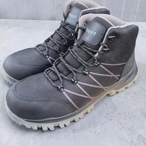 Puede incluir: Un par de botas al tobillo negras Propét con puntera texturizada, panel lateral de malla con cordones cruzados, ojales metálicos, forro interior gris y una suela de goma gruesa de color gris claro con tacos para mayor tracción.