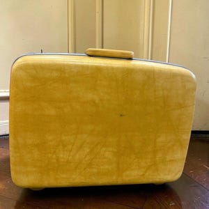 May include: A rectangular, mustard yellow hard-shell suitcase with a textured surface. The suitcase features a top-mounted handle and rounded corners, presenting a solid, structured design for travel storage and organization.