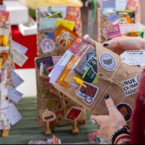 May include: A brown paper package tied with twine, containing books and treats. Stickers include a bear, a telephone with text "What's your favorite scary movie?", and "True Crime Fanatic". A tag reads "Mystery/Thriller". A yellow pen is tucked inside.