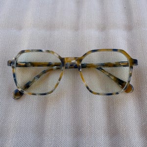 May include: A pair of eyeglasses with a tortoiseshell pattern. The frames are a mix of brown, yellow, and blue, with a geometric shape. The lenses are clear. The glasses are resting on a neutral-colored surface.
