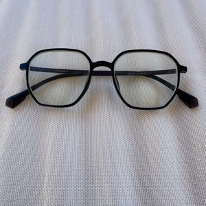 May include: Black, geometric eyeglasses with clear lenses. The frames have a matte finish and a subtle, angular design. The temples are dark and extend to the ear. The glasses are set against a white, textured background.