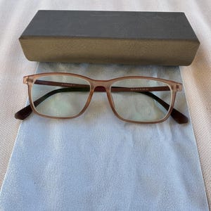 May include: Rectangular eyeglasses with a brown frame and dark temples rest on a light blue microfiber cloth. A dark gray rectangular glasses case sits above the glasses. The glasses have clear lenses.