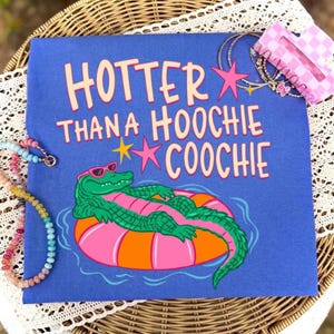 May include: Blue t-shirt with the text "Hotter Than A Hoochie Coochie" in pink letters. The shirt features a cartoon alligator wearing sunglasses and floating on a pink and orange inner tube. Accessories include a pink hair clip and bracelets.