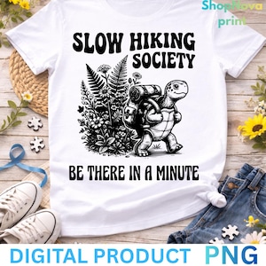Può includere: T-shirt bianca con grafica nera di una tartaruga con zaino, che cammina vicino a fiori. Il testo recita "SLOW HIKING SOCIETY" e "BE THERE IN A MINUTE". La maglietta è esposta su una superficie di legno con altri oggetti.