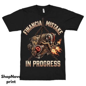 Pode incluir: Camiseta preta com uma ilustração detalhada de um motor, chamas e o texto "FINANCIAL MISTAKE IN PROGRESS". O motor está suspenso por correntes, com uma luz vermelha brilhante do motor.