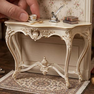 Może przedstawiać: Miniaturowa konsola w kolorze kremowym ze złotymi akcentami. Na stole stoi maleńka filiżanka ze spodkiem, pióro i kałamarz oraz miniaturowe książki. Stół stoi na wzorzystym dywanie, z ręką umieszczającą filiżankę.