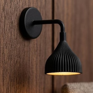 Puede incluir: Una lámpara negra montada en la pared con una pantalla acanalada está fijada a una pared de madera. La lámpara está encima de una pequeña mesa con un libro abierto, gafas y un libro cerrado. La escena está iluminada por la cálida luz de la lámpara.