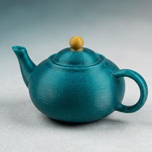 May include: A small, round teapot in a metallic teal finish with a curved spout and a circular handle. The lid features a small, spherical knob in a contrasting matte gold color. The surface of the teapot displays fine, horizontal 3D printed lines.