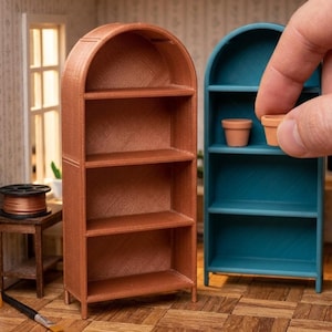May include: Two miniature arched shelving units, one in a metallic copper finish and one in a matte teal finish. The teal unit features three horizontal shelves and contains two small terracotta-colored pots, one being placed by a finger.