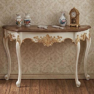 以下が含まれることがあります： 曲線的な天板と装飾的なディテールが特徴の、白と金のコンソールテーブル。テーブルには、花柄の小さな花瓶3つ、小さな時計、装飾品が置かれています。背景は模様入りの壁紙です。