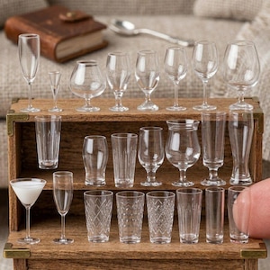Könnte beinhalten: Eine Sammlung von Miniatur-Trinkgläsern aus klarem Glas, darunter Weingläser, Sektflöten und verschiedene Becher, die auf einer Holzkiste ausgestellt sind. Das Sortiment zeigt verschiedene Formen und Größen, perfekt für ein Puppenhaus oder eine Miniatursammlung.