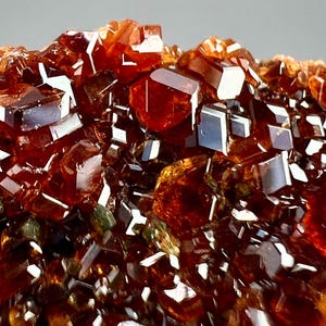 Può includere: Un grappolo di cristalli di granato color arancione intenso e marrone rossastro con una finitura traslucida e lucida. Il campione minerale presenta numerose formazioni cristalline geometriche sfaccettate che catturano la luce.