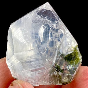 May include: A translucent, multifaceted crystal specimen featuring a clear, colorless body with distinct internal striations and a prominent cluster of vibrant green mineral inclusions concentrated along the lower right edge of the stone.