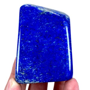 May include: A polished, rectangular lapis lazuli stone. The deep blue stone has a glossy finish and is flecked with small, gold-colored inclusions. The stone is held in a hand, showcasing its vibrant color and natural beauty. The shape is slightly trapezoidal.