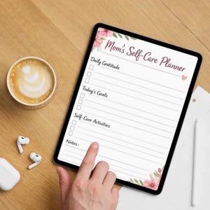 May include: A digital tablet displaying a "Mom's Self-Care Planner" with sections for daily gratitude, goals, and activities. The tablet is on a wooden surface, next to a cup of coffee and wireless earbuds. A hand is touching the screen.