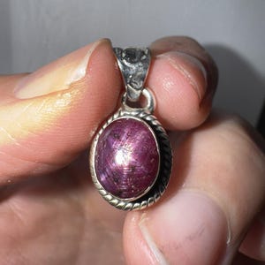 May include: Oval pendant featuring a deep purple gemstone. The gemstone is set in a silver-coloured metal frame with a twisted rope design. The pendant has a small loop at the top for a chain, suitable for a necklace.