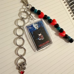 May include: A keychain featuring a clear plastic Hellraiser movie image, connected to a silver chain and a beaded strand. The beaded strand has black, teal, and red heart-shaped beads. A small red heart charm hangs from the chain.
