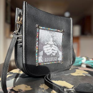 May include: Black crossbody bag with a textured finish. The bag features a rectangular patch with the text "DANZIG II LUCIFUGE" and a graphic. The patch is framed with colorful rhinestones. The bag has a long, adjustable strap.