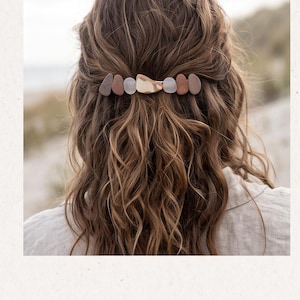 May include: A hair clip featuring a row of small, smooth, pebble-shaped elements in shades of brown, white, and translucent gray, centered with a larger, irregular, metallic gold-toned piece, secured in wavy, light brown hair.