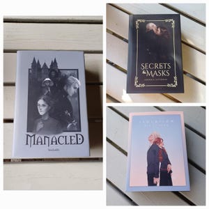 Peut inclure: Trois livres reliés avec des couvertures illustrées. Le livre de gauche est gris clair et intitulé Manacled par SenLinYu. Le livre en haut à droite est noir et intitulé Secrets & Masks par Emerald Slytherin. Le livre en bas à droite est bleu clair et intitulé Isolation.