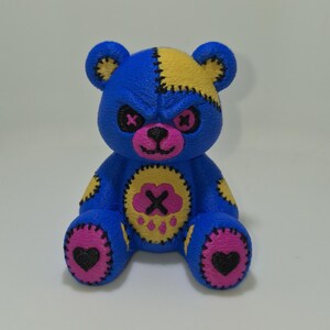 May include: A blue teddy bear figurine featuring yellow patches on the head and limbs, pink button-style eyes with black X stitching, a pink nose, and a central yellow belly patch with a pink cloud and black X design, plus pink paw pads.