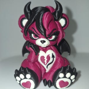 May include: A 3D printed bear figurine in magenta and black. The bear has black horns, black bat wings, and a white heart on its chest with a broken heart and arrow design. It features black paw pads with magenta hearts and a stern facial expression.