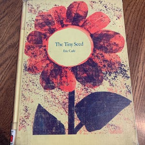 Puede incluir: Un libro infantil titulado "The Tiny Seed" de Eric Carle. La portada presenta una gran flor estilizada en tonos rojos y morados sobre un fondo amarillo. El libro tiene un aspecto desgastado.