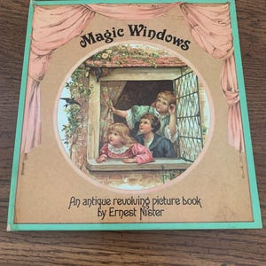 May include: An antique children's book titled "Magic Windows" by Ernest Nister. The cover shows a circular illustration of children looking out a window, framed by pink curtains and a green border. The book is on a wooden surface.