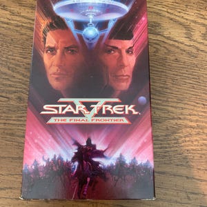 Peut inclure: Une boîte de cassette VHS rectangulaire pour Star Trek V: The Final Frontier. La couverture présente un fond spatial rouge et bleu avec l'USS Enterprise, les portraits de deux personnages et la silhouette d'une figure à cheval en bas.