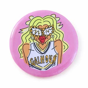 Puede incluir: Un botón redondo rosa con una ilustración de dibujos animados de una mujer con cabello rubio, labios rojos grandes y gafas. La mujer lleva una camiseta blanca y amarilla con la palabra "CALHOUN".