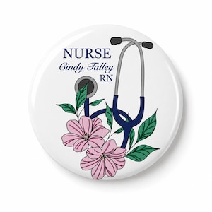Könnte beinhalten: Ein runder weißer Ansteckbutton mit einer dunkelblauen Stethoskop-Grafik, die mit rosa Blüten und grünen Blättern verziert ist. Der Text lautet NURSE, Cindy Talley und RN in dunkelblauer Schrift. Das Design ist auf weißem Grund zentriert.