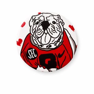 May include: A round white button featuring a bulldog illustration in black and white, wearing a red jersey with the text "SEC" and the number 2. Red heart shapes are scattered around the edge.