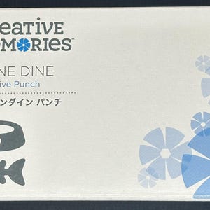 Puede incluir: Una caja blanca con el texto "creative memories" y "FELINE DINE Decorative Punch". La caja presenta un cuenco para gatos y un gráfico de espina de pescado. Los diseños de flores azules están en la esquina. También hay texto en japonés.