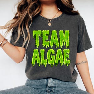 May include: A charcoal gray short-sleeve t-shirt featuring the text "TEAM ALGAE" printed in a bright, dripping lime green font. The graphic design has a liquid, melting appearance, and the shirt has a faded, washed-out fabric texture.