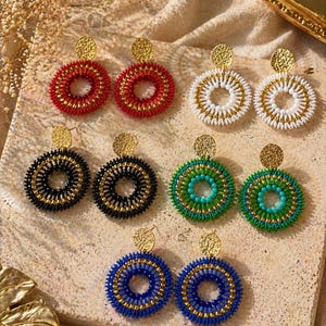 May include: A selection of beaded hoop earrings in different colours. The earrings have a circular design with a smaller inner circle. Colours include red, white, black, green, and blue, with gold-coloured accents. The earrings are presented on a neutral surface.