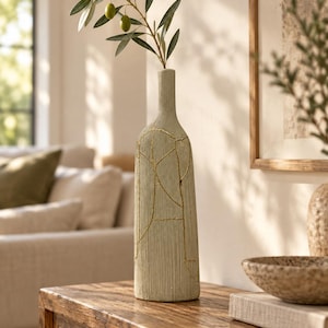 May include: A tall, cylindrical, light green vase with a textured, ribbed surface and thin, irregular gold-colored lines forming a map-like pattern. A single green olive branch with three small green olives is placed inside the narrow neck.
