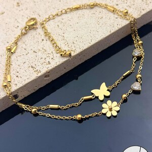 Puede incluir: Tobillera dorada con dos cadenas delicadas. Una cadena tiene un dije de mariposa, mientras que la otra tiene dijes de flores y corazones. La tobillera está adornada con pequeñas cuentas y está diseñada para usarse alrededor del tobillo.
