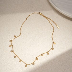 May include: A delicate gold-colored necklace with a series of small, teardrop-shaped pendants and round, clear gemstones. The necklace is displayed on a neutral surface, showcasing its elegant design. The necklace is a jewelry item.