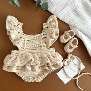 May include: A beige crochet romper featuring ruffled shoulder straps and a ruffled waistline, accompanied by a pair of matching crochet Mary Jane shoes and a crochet headband with a bow, displayed with a product tag labeled Tiny Baby Collections.