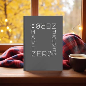 May include: A gray journal with the words "HAVE ZERO THOUGHTS" in white lettering. The journal is on a wooden surface next to a cup of coffee and a plaid blanket. The background shows a window with fall foliage.