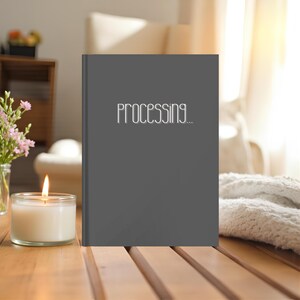 May include: A gray book with the word "PROCESSING..." in white lettering. A lit candle in a glass jar sits on a wooden surface. Pink flowers in a glass vase are also on the table.